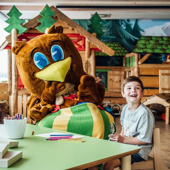 Ein Junge lacht neben einem Maskottchen in einem Spielzimmer mit bunten Wänden und Holzmöbeln.