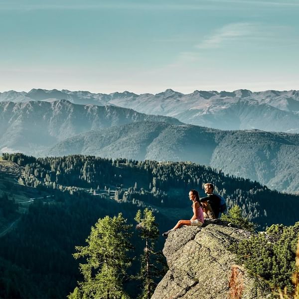 Zwei Wanderer sitzen auf einem Felsvorsprung und genießen die Aussicht auf bewaldete Berge und Himmel.
