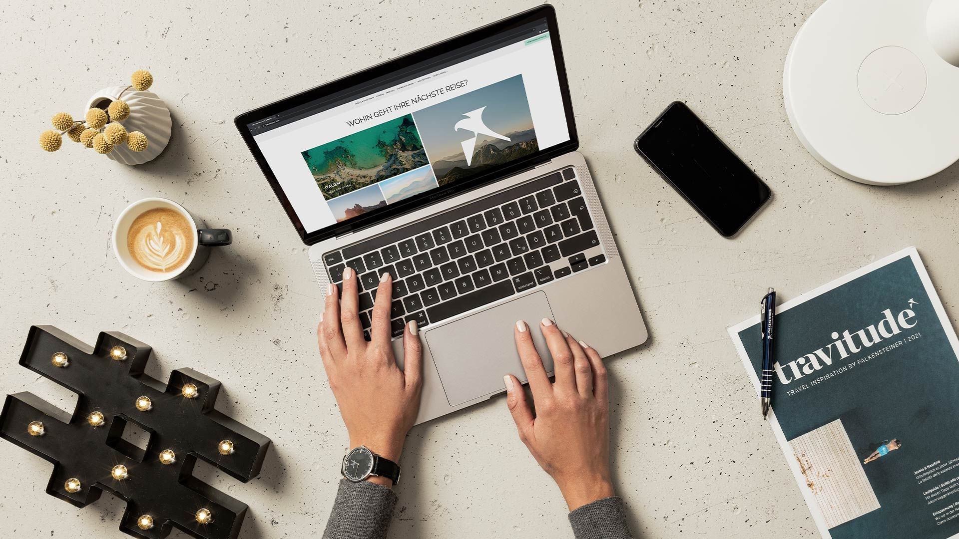 Hände tippen auf Laptoptastatur, umgeben von Smartphone, Kaffee, Magazin und Notizbuch auf Tisch.