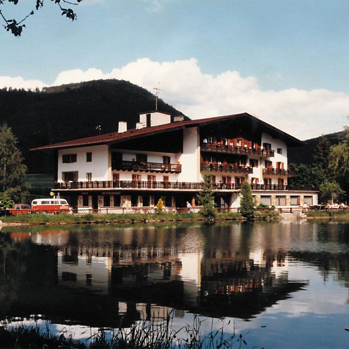 Weißes Gebäude mit mehreren Balkonen, das sich an einem See oder Teich spiegelt.