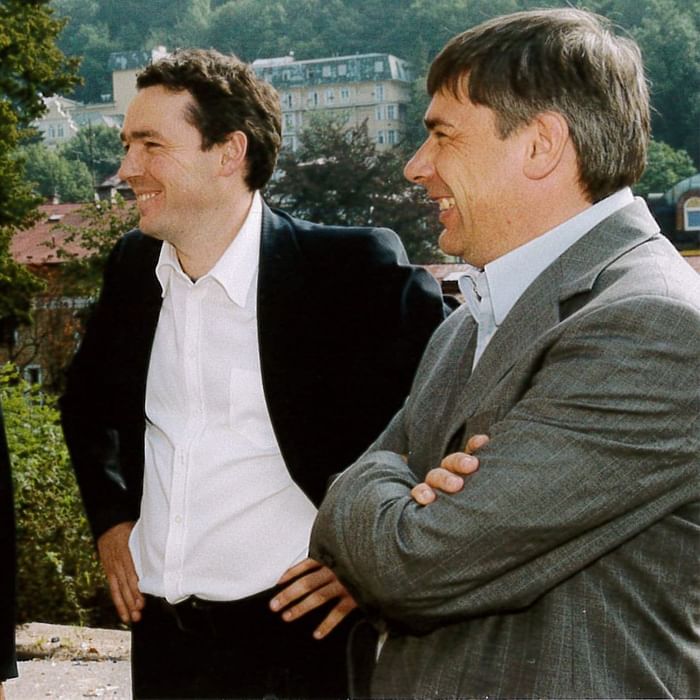Zwei Männer in Anzügen lächeln und unterhalten sich vor einem Hintergrund mit Bäumen und Gebäuden.