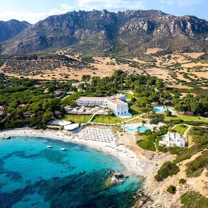 Luftaufnahme eines Resorts mit Gebäuden, Pool, Strand und Berglandschaft im Hintergrund.
