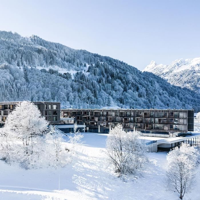 Modernes Gebäudekomplex mit vielen Fenstern inmitten von schneebedeckten Bergen und Bäumen.