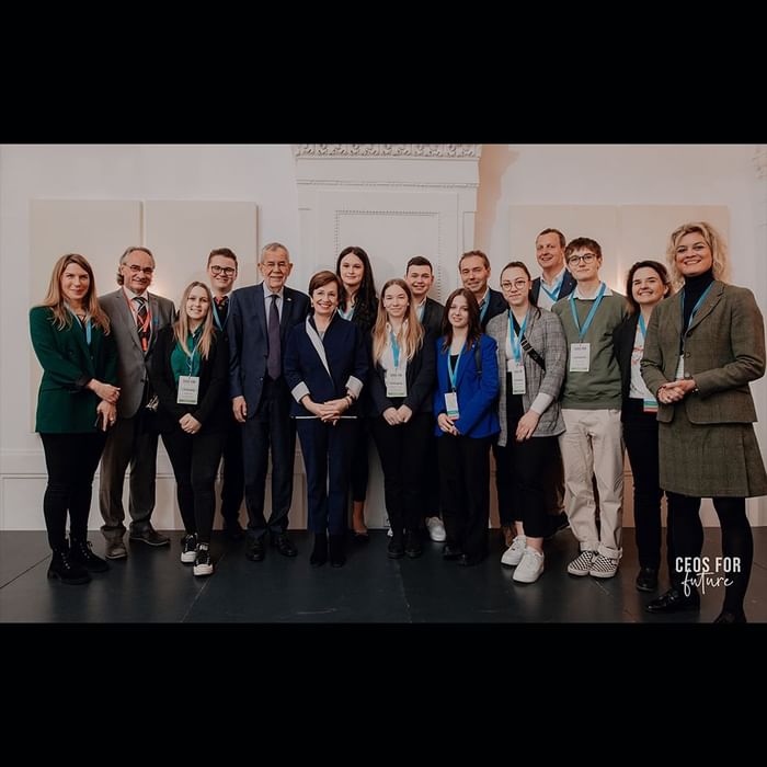Gruppe von Personen in formeller Kleidung, die nebeneinander vor einer weißen Wand stehen und lächeln.