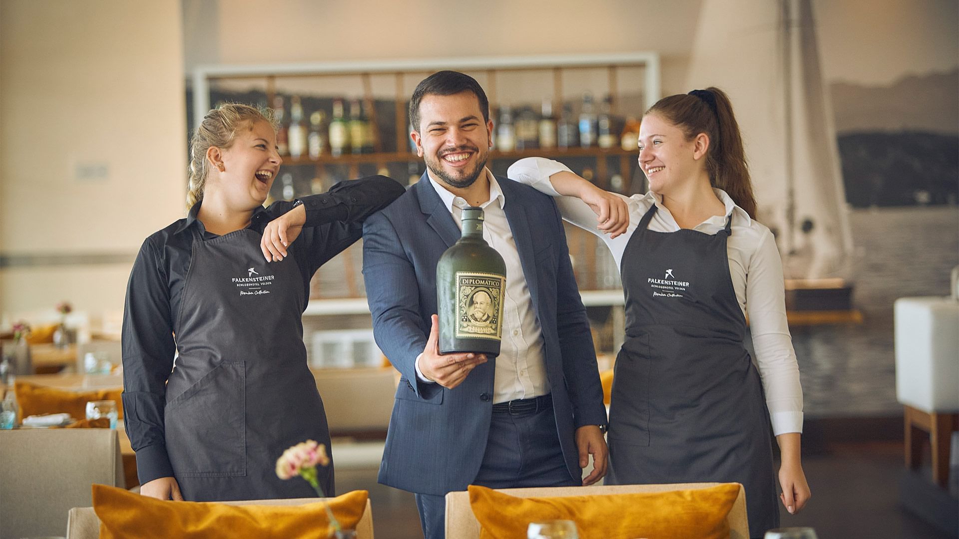 Drei Personen in einem Restaurant, zwei tragen identische schwarze Schürzen und der Mann hält eine große Flasche.