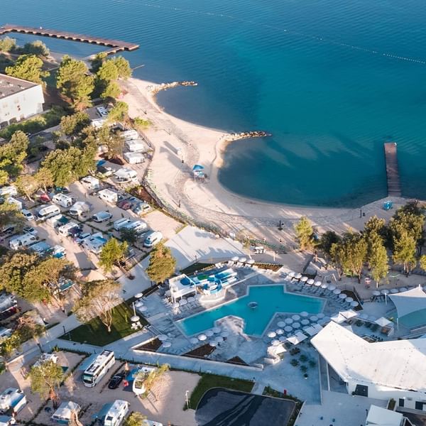 Luftaufnahme eines Resorts mit Pool, Strand, Gebäuden, geparkten Fahrzeugen und umliegenden Bäumen.