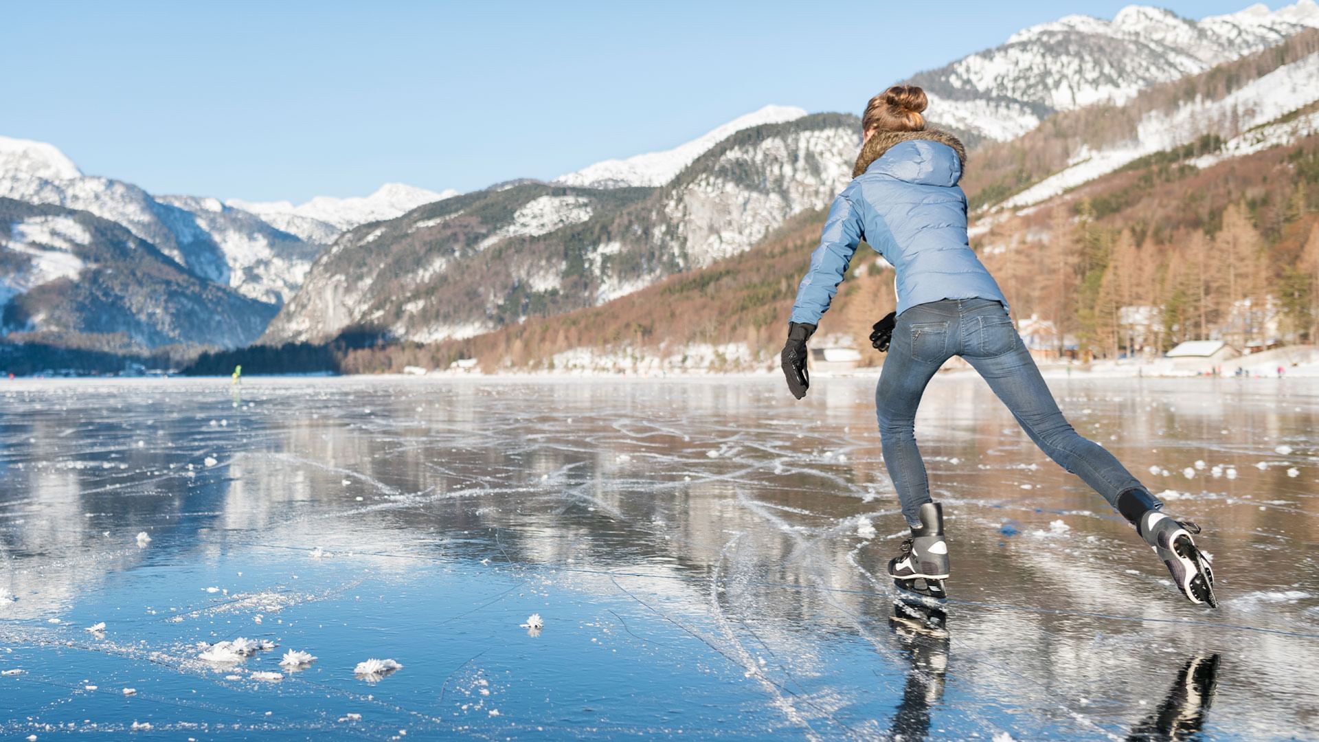 Frau im blauen Mantel und Jeans eisläuft auf gefrorenem See mit verschneiten Bergen im Hintergrund.