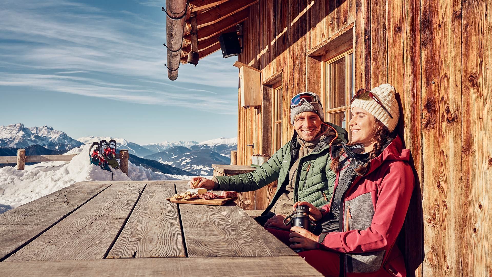 Zwei Personen sitzen an einem Holztisch vor einem Holzgebäude und genießen die Aussicht.