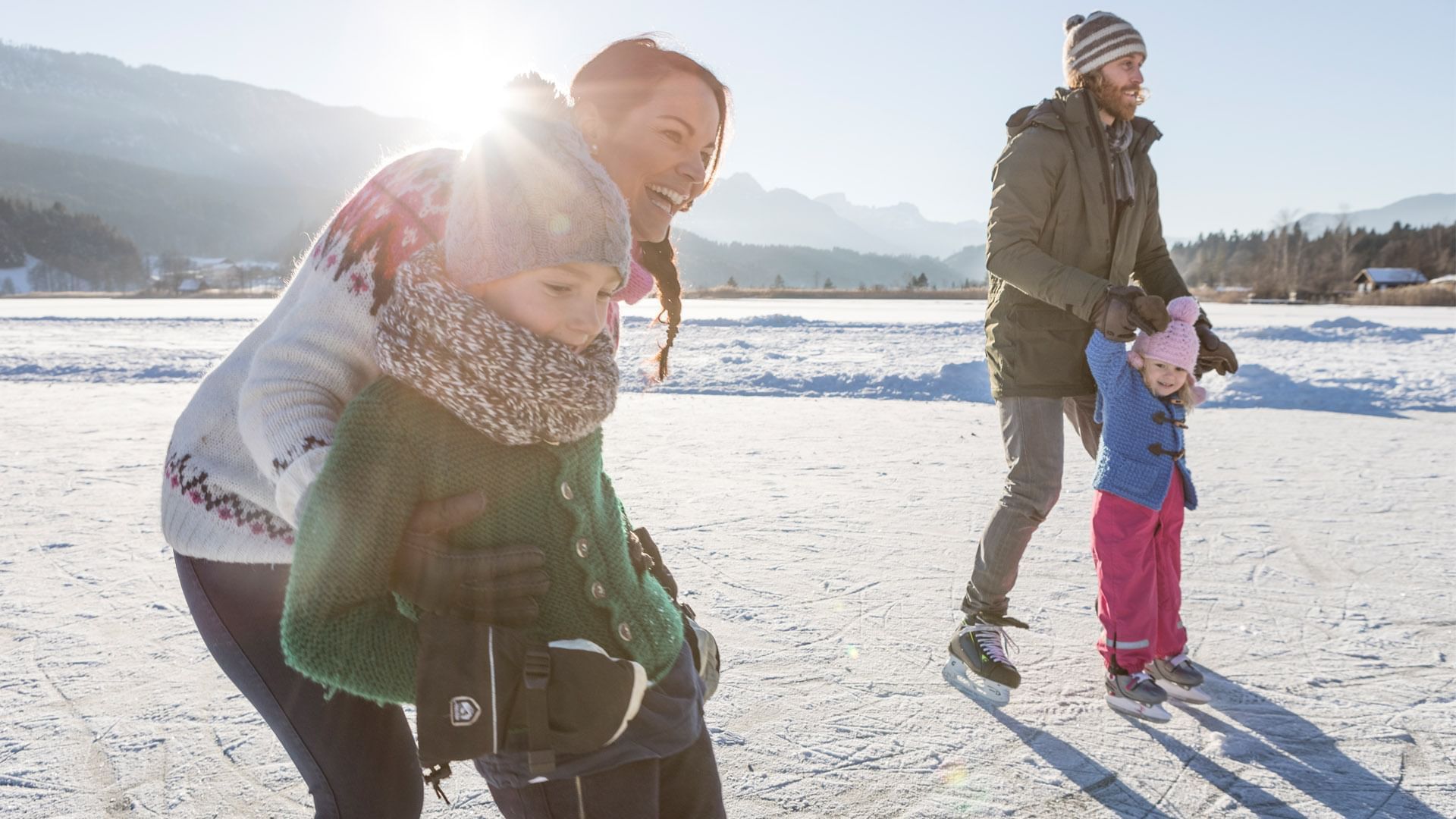 Familie beim Eislaufen auf einem verschneiten See mit Bergen im Hintergrund.