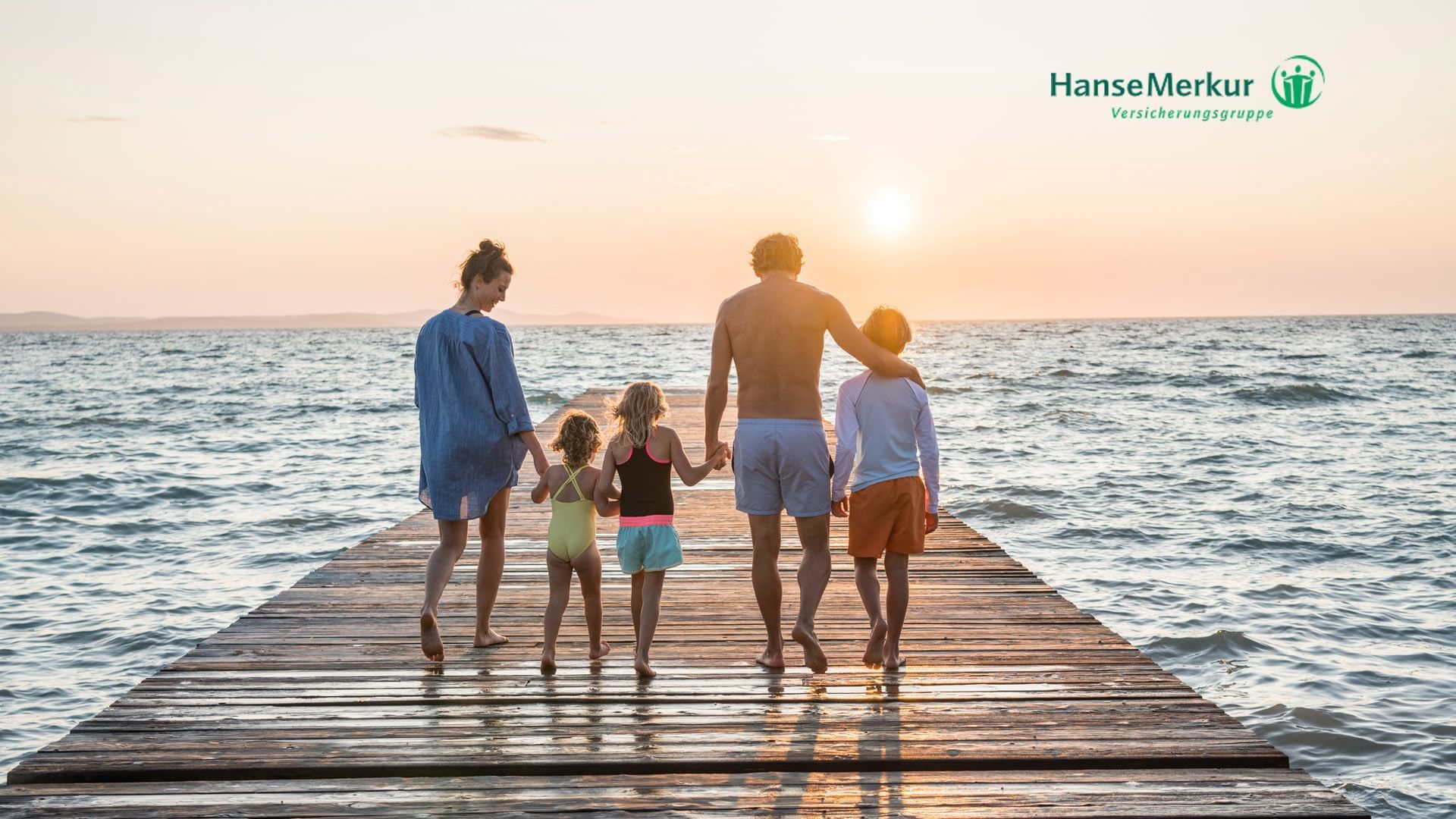 Familie auf Holzsteg im Meer bei Sonnenuntergang, Logo von HanseMerkur Versicherungsgruppe im oberen rechten Eck.