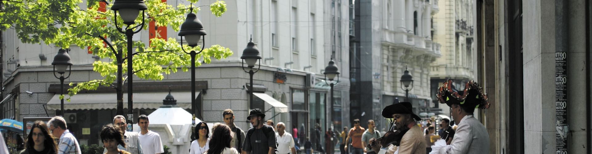 Zwei Musiker in bunten Hüten spielen für Passanten auf einer belebten Stadtstraße mit Bäumen und Gebäuden.