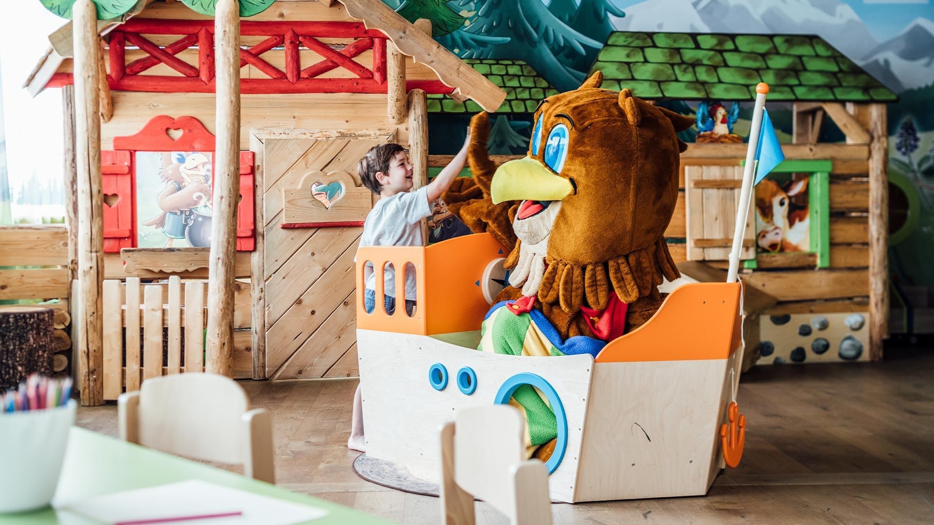 Kind spielt mit einem Eulen-Maskottchen in einem Holzspielhaus in einem Raum mit Waldmotiven.