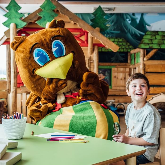 Junge sitzt mit Stiften und Lupe an Tisch, neben braunem Maskottchen mit gelbem Schnabel und buntem Körper.