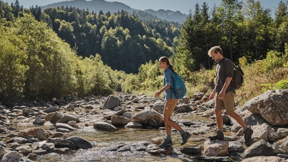Zwei Wanderer mit Rucksäcken überqueren einen felsigen Bach in einer bewaldeten Landschaft.