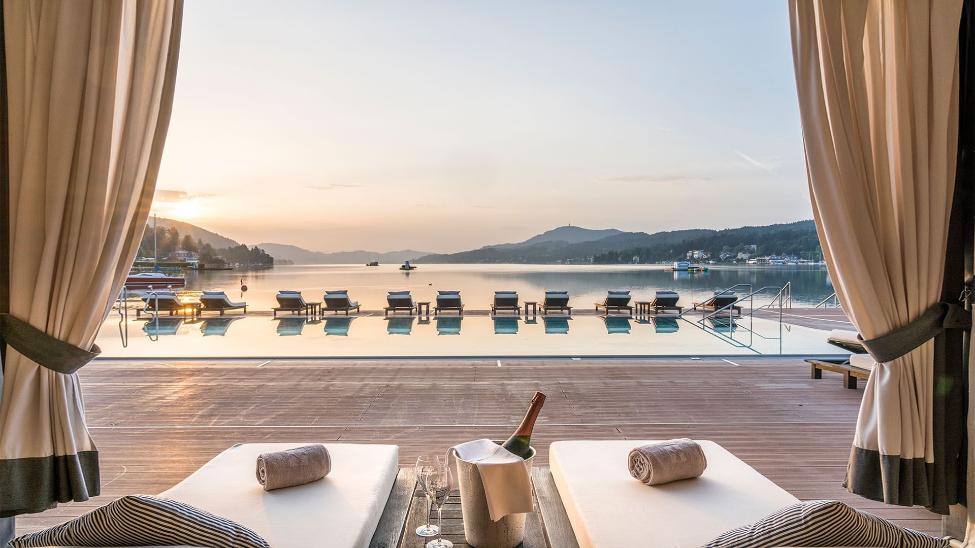 Sonnenuntergangsblick auf einen Pool mit Liegen, begleitet von Champagner und umgeben von Wasser und Bergen.