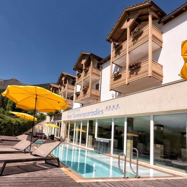 Sonnenparadies Hotel mit Pool, Liegen, gelben Schirmen und blühenden Balkonen an sonnigem Tag.