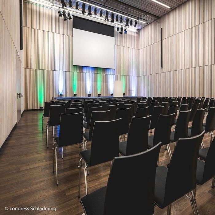 Leerer Konferenzraum mit schwarzen Stühlen, Holzboden, Leinwand und grünen Lichtern an der Wand.