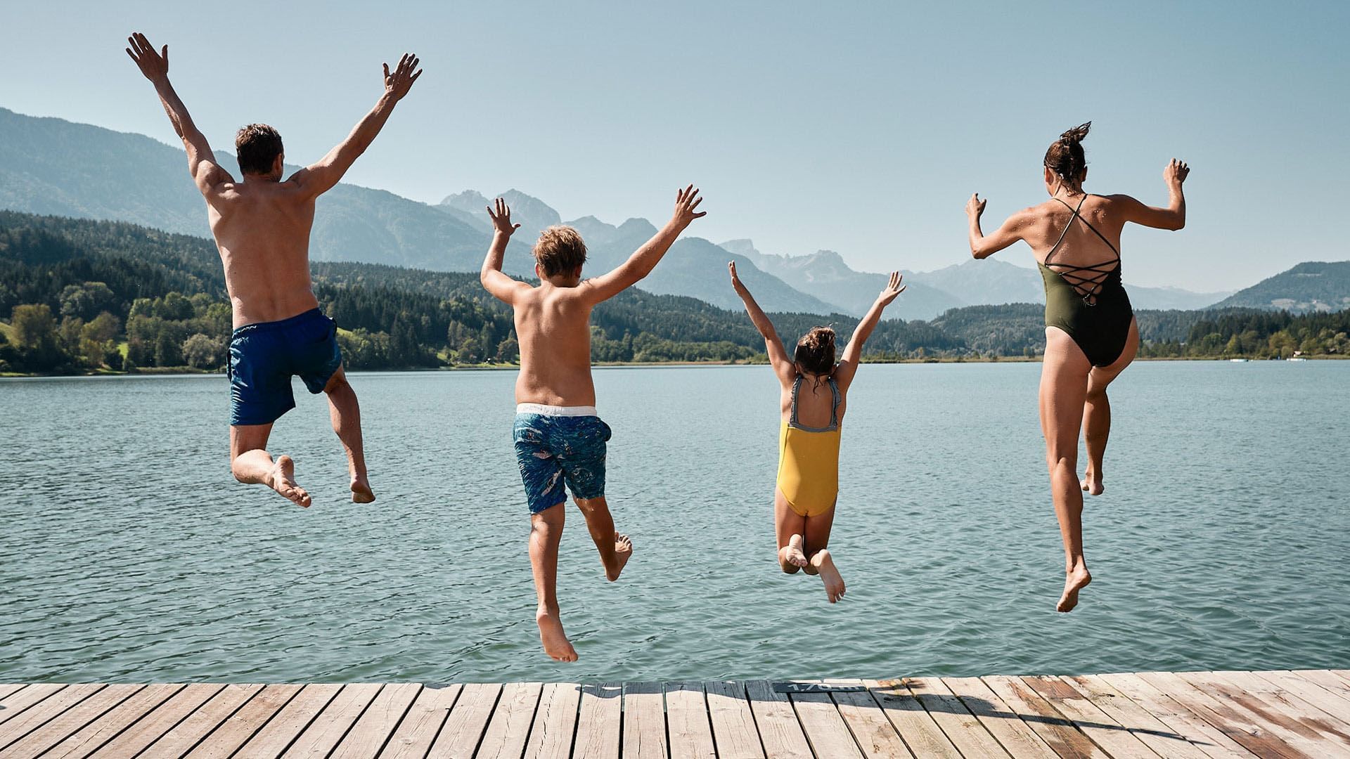 Vier Personen springen gemeinsam von einer Holzplattform in einen See mit Bergkulisse im Hintergrund.
