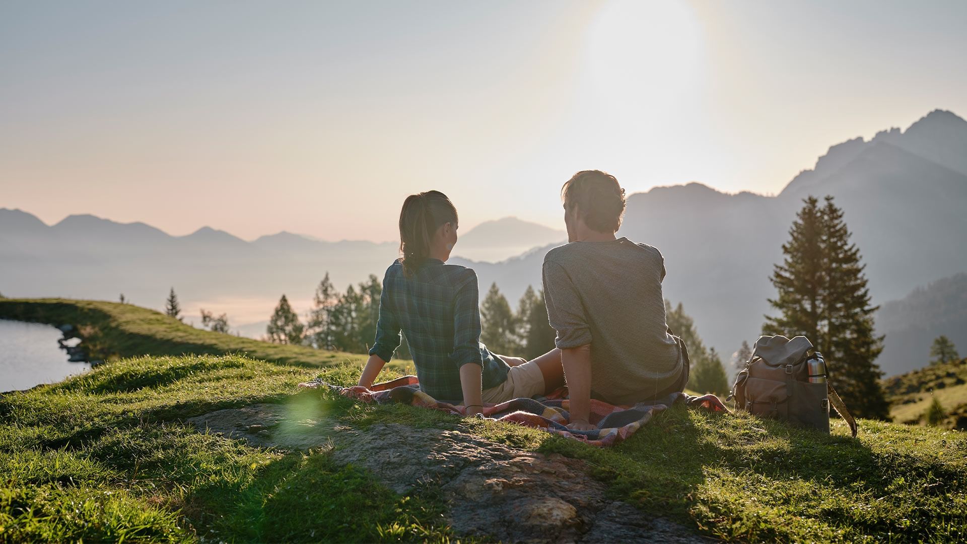 Zwei Personen sitzen auf einer Decke auf einer Wiese und betrachten den Sonnenuntergang über einem Bergsee.
