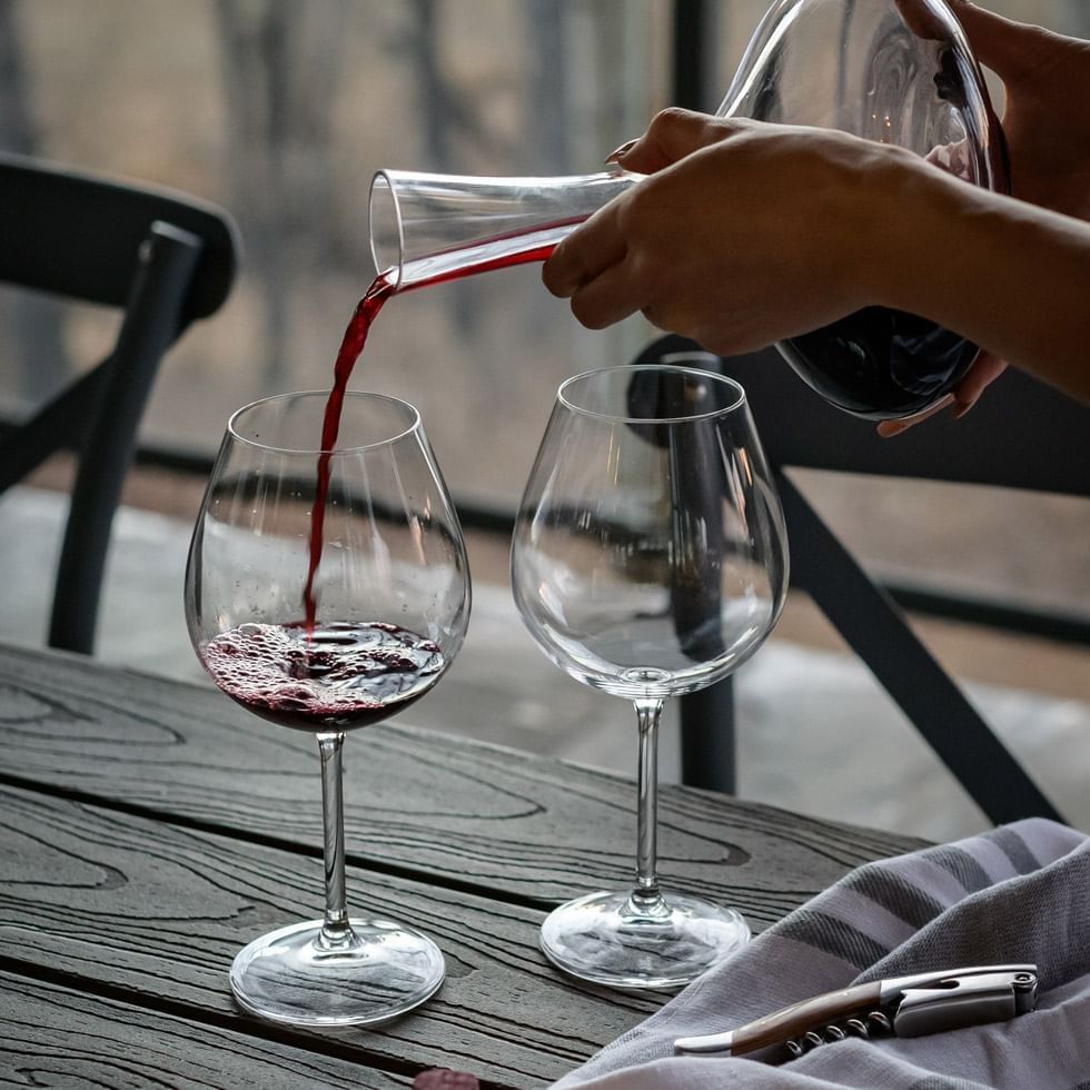Eine Person gießt Rotwein aus einer Karaffe in ein Glas auf einem hölzernen Tisch.