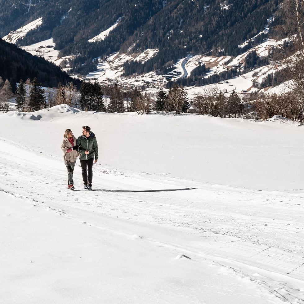 Zwei Personen gehen Hand in Hand über eine schneebedeckte Landschaft mit Bergen im Hintergrund.