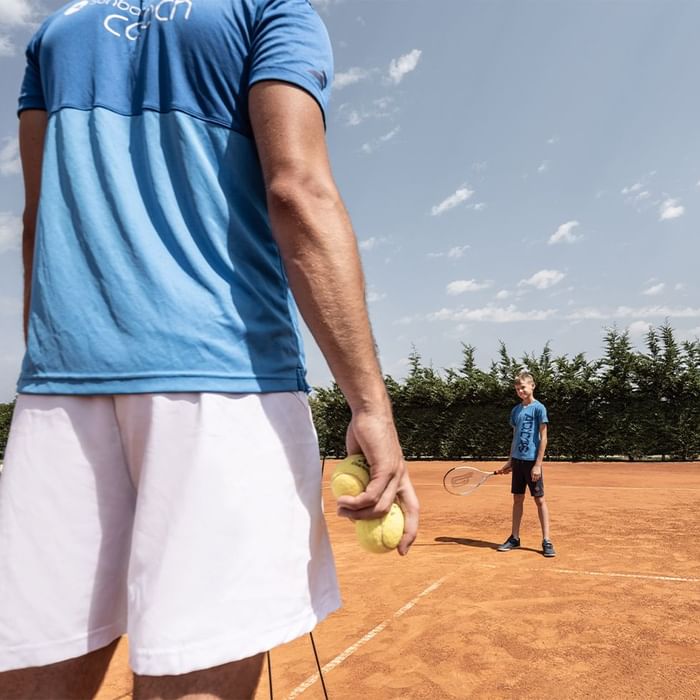 Trainer und Schüler auf einem Tennisplatz mit Schlägern und Bällen, Bäume im Hintergrund.