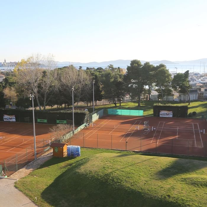 Tennisplätze mit Zaun und Bäumen, umgeben von Gras und Blick auf die Küste.