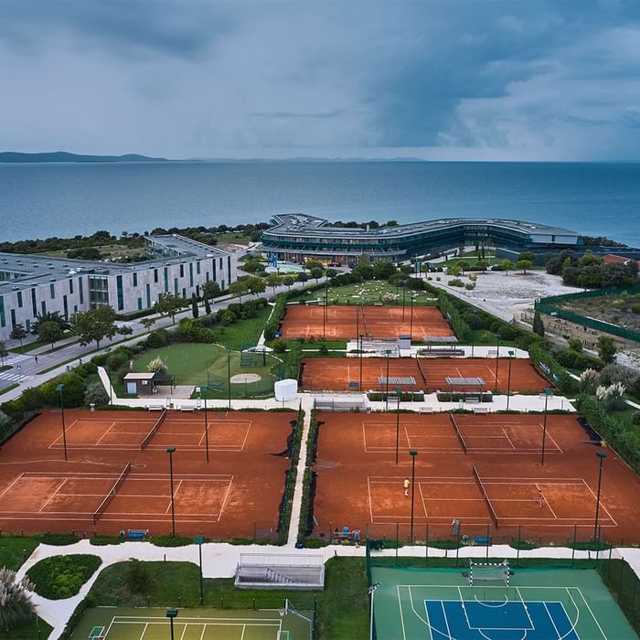 Luftaufnahme eines Sportkomplexes mit mehreren Tennisplätzen, einem Basketballplatz und Gebäuden am Meer.
