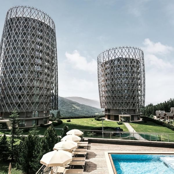 Zwei moderne zylindrische Gebäude neben einem Pool und Liegestühlen, umgeben von Bäumen und Bergen.