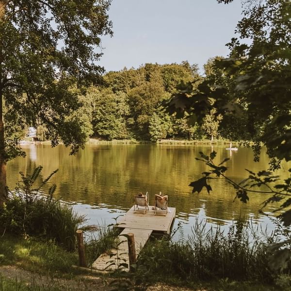Zwei Personen sitzen auf Liegen auf einer hölzernen Plattform neben einem See, umgeben von Bäumen.