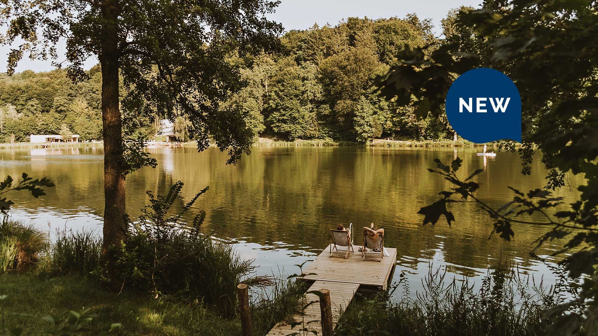 Zwei Personen sitzen auf Liegen auf einer Holzplattform am Ufer eines Sees, umgeben von Bäumen.