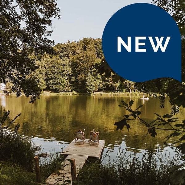 Zwei Personen sitzen auf Stühlen auf einer Plattform am See, umgeben von Bäumen und einem 