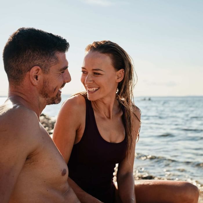 Mann und Frau lächeln sich am Strand zu, Wasser im Hintergrund.