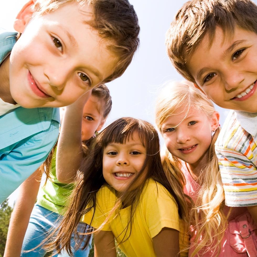 Fünf Kinder stehen nahe beieinander und lächeln in die Kamera, wobei ein weiteres Kind im Hintergrund steht.