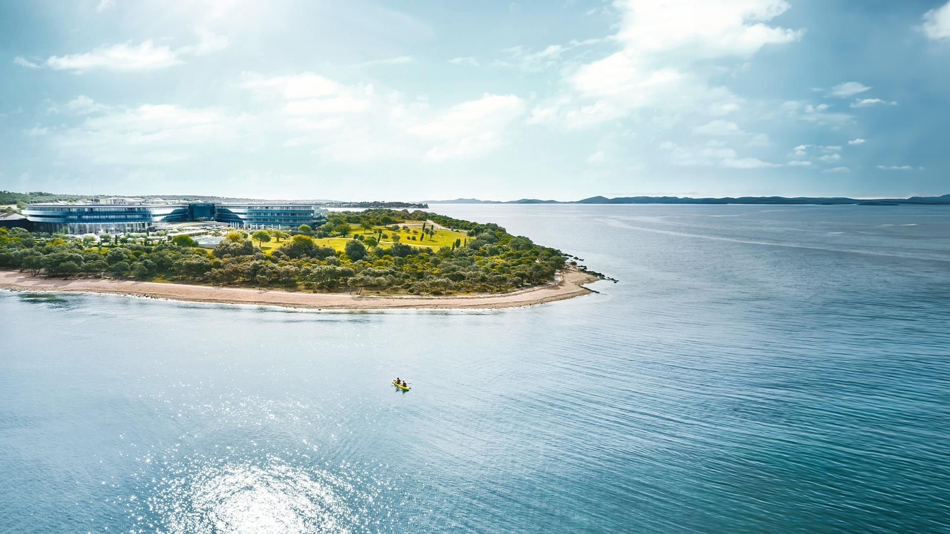 Vista aerea di un'isola con edificio moderno e kayak nell'acqua blu sotto un cielo nuvoloso.