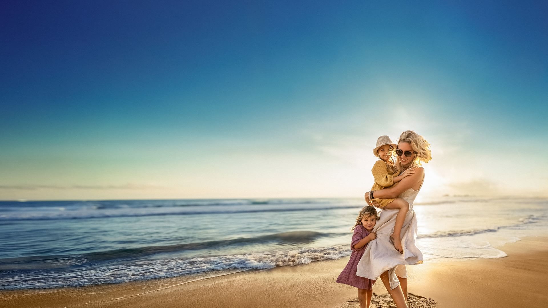 Mutter mit zwei Kindern am Strand, einer auf dem Arm, bei Sonnenuntergang.