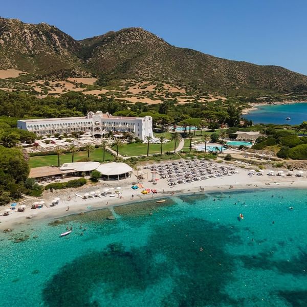 Luftaufnahme eines Resorts mit Pool, Strand und blauem Meer, umgeben von Hügeln und Vegetation.