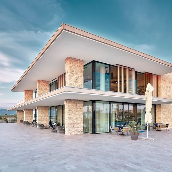 Moderne Villa mit Glasfassade und Terrasse, umgeben von Stühlen, Tischen und einer weißen Sonnenschirm.