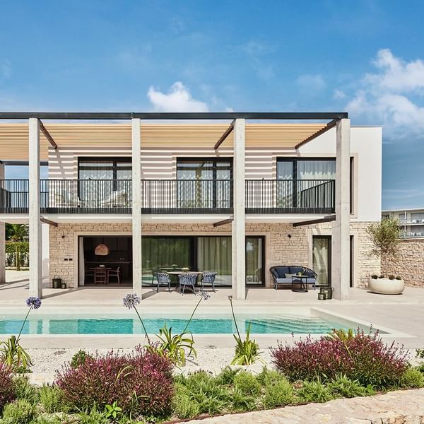 Modernes Haus mit Pool und Terrasse, umgeben von Pflanzen unter einem blauen Himmel mit Wolken.