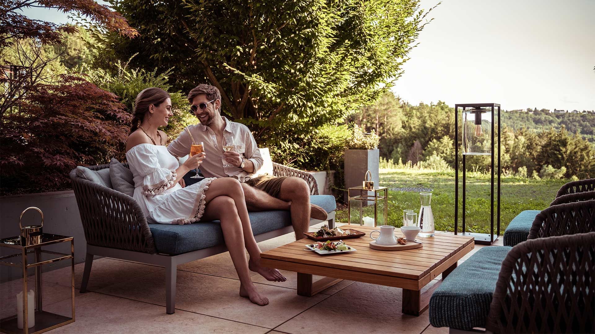 Coppia seduta su divano da giardino, sorridente e con bicchieri di vino, vicino a tavolo con cibo.