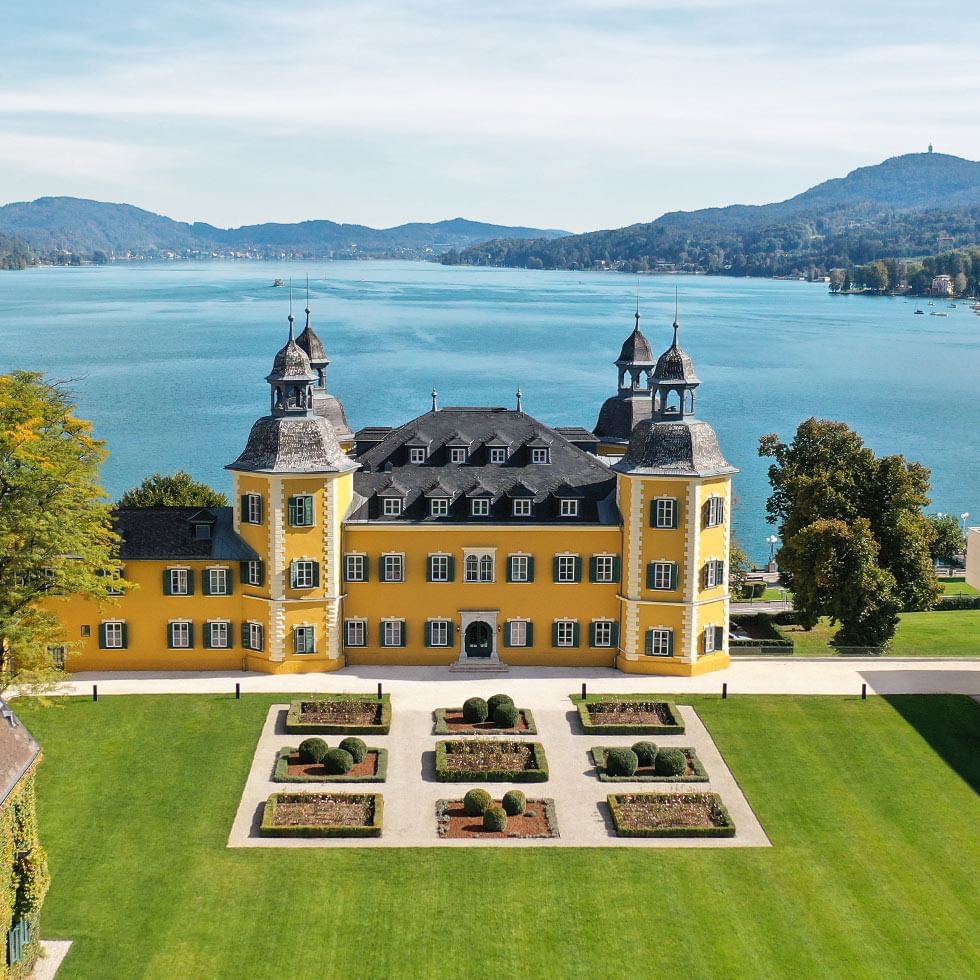 Gelbes Schloss mit Garten vor einem See und Hügeln im Hintergrund, aufgenommen aus der Vogelperspektive.