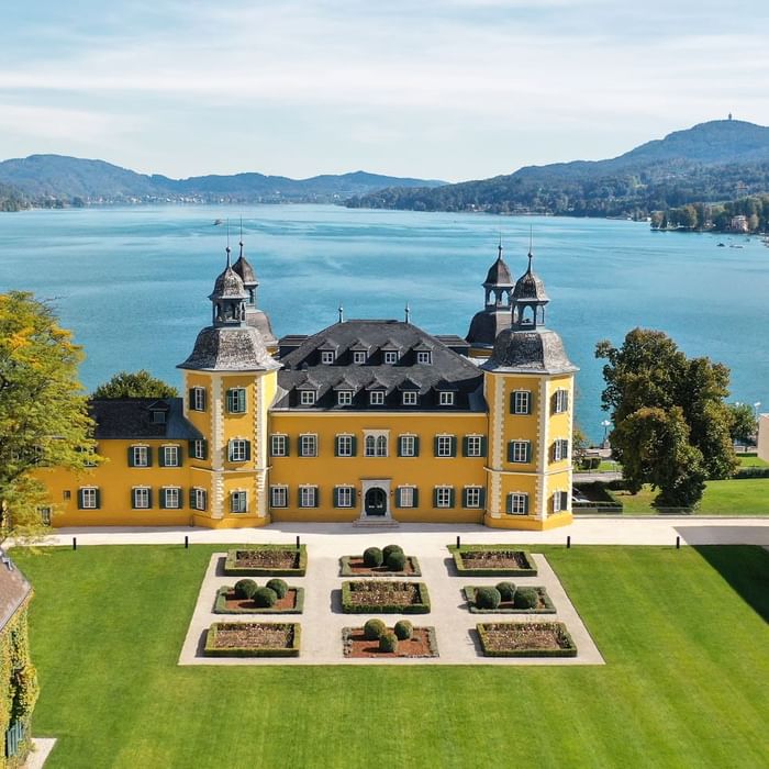 Gelbes Schloss mit Garten vor einem See und Hügeln im Hintergrund, aufgenommen aus der Vogelperspektive.