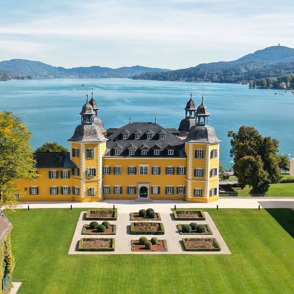 Gelbes Schloss mit Garten vor einem See und Hügeln im Hintergrund, aufgenommen aus der Vogelperspektive.