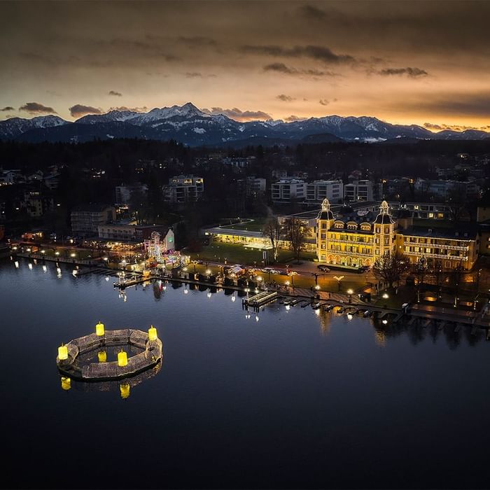 Luftaufnahme einer Stadt mit beleuchtetem See und einer beleuchteten, kreisförmigen Struktur im Wasser.