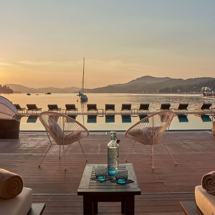 Terrasse mit zwei Sesseln und einem Tisch am Pool, im Hintergrund ein See und eine untergehende Sonne.