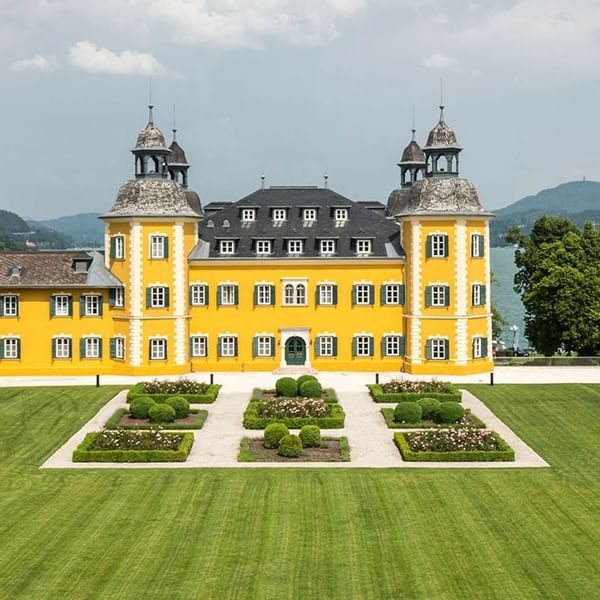 Gelbes Schloss mit zwei Türmen und Garten vor einem See und Bergen im Hintergrund.