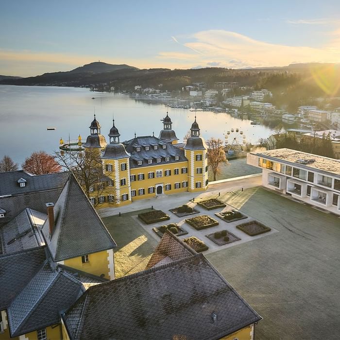Luftaufnahme eines prächtigen Schlosses am See mit umliegenden Gebäuden und einer Stadt in der Ferne.