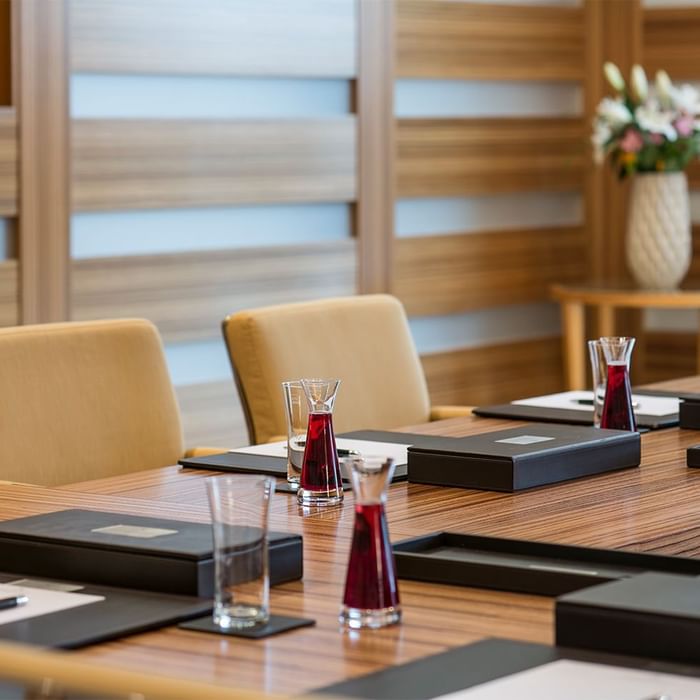 Konferenztisch mit zwei Stühlen, Gläsern, Ordnern und einer Vase mit Blumen im Hintergrund.