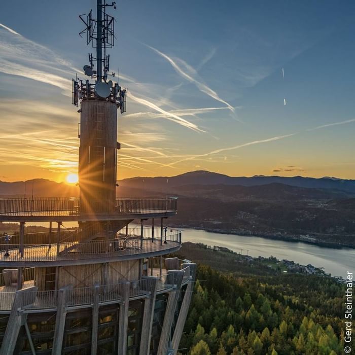 Sonnenuntergang hinter einem Fernmeldeturm mit Blick auf einen See und bewaldete Hügel im Hintergrund.