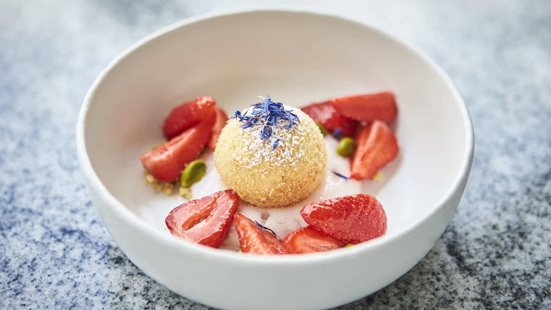 Weiße Schüssel mit Erdbeeren, einem runden Dessert und blauen Blüten auf einer steinernen Oberfläche.